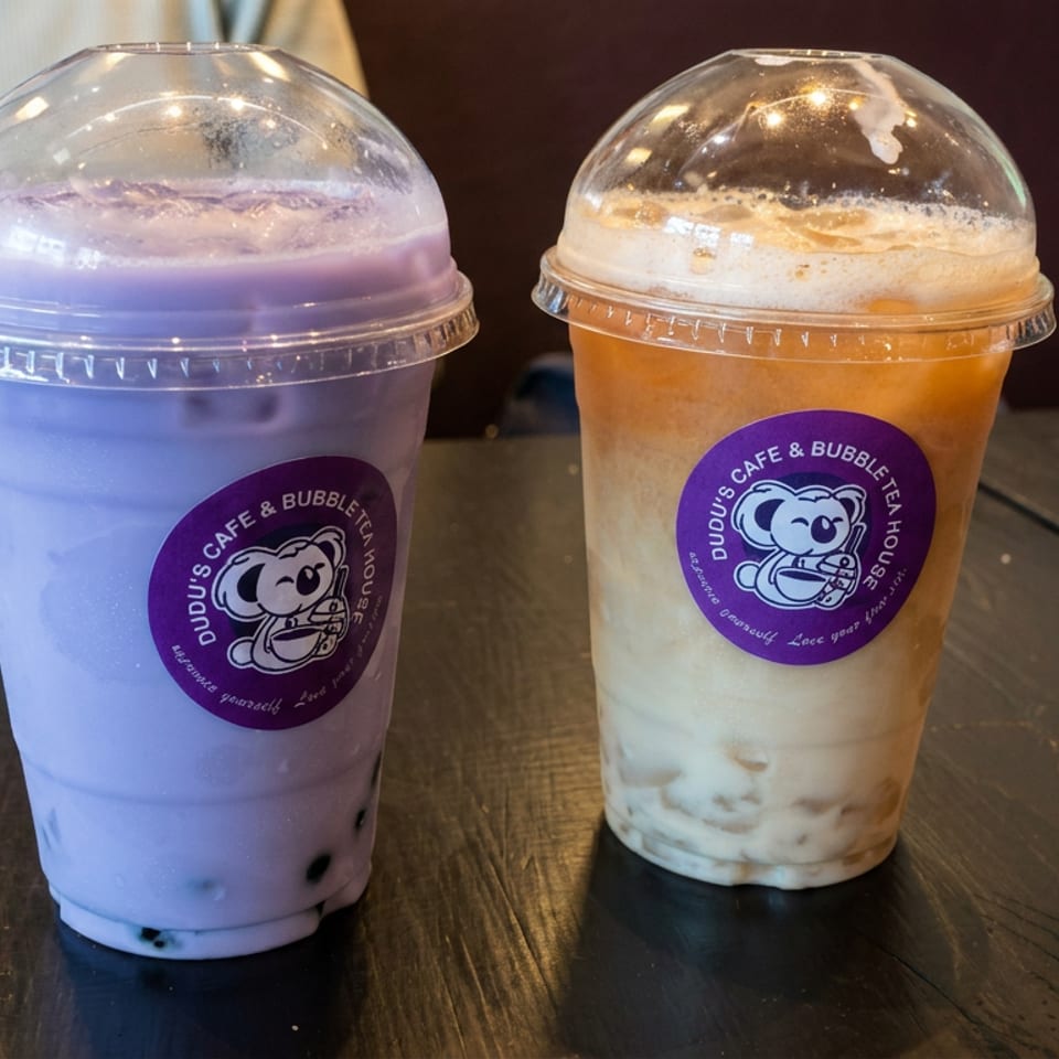 Two colorful bubble tea drinks with tapioca pearls and domed lids sit on a dark wooden table at Udou’s Cafe & Bubble Tea House.