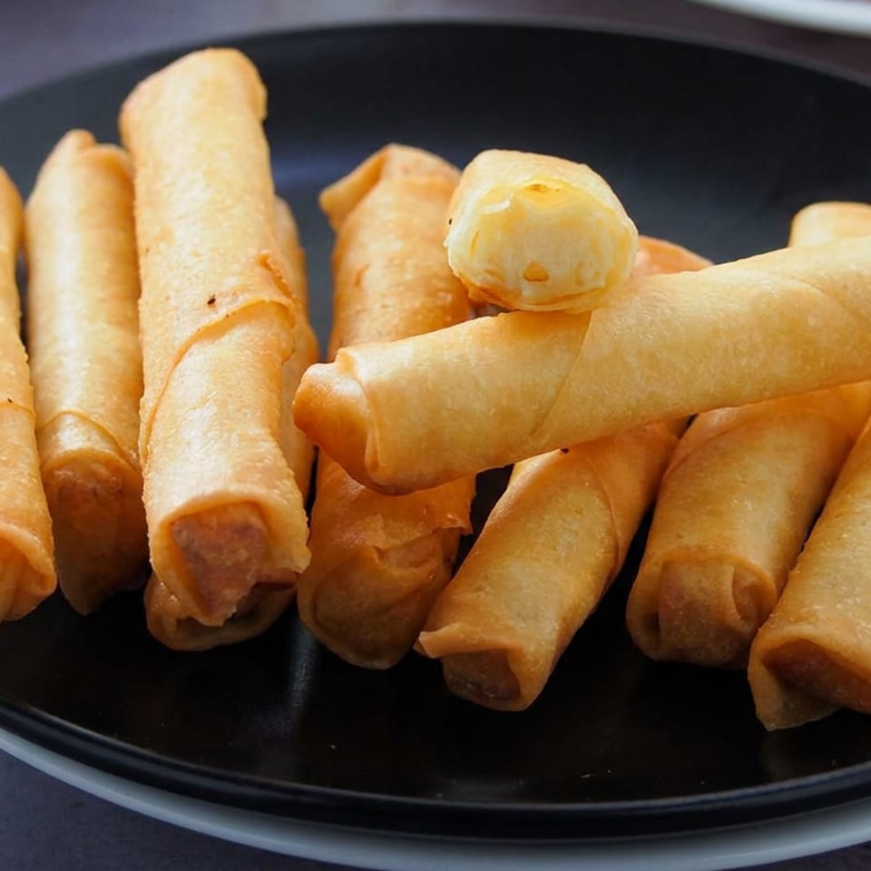 A black plate holds a pile of golden-brown fried spring rolls, with one topped by a small dollop of sauce.