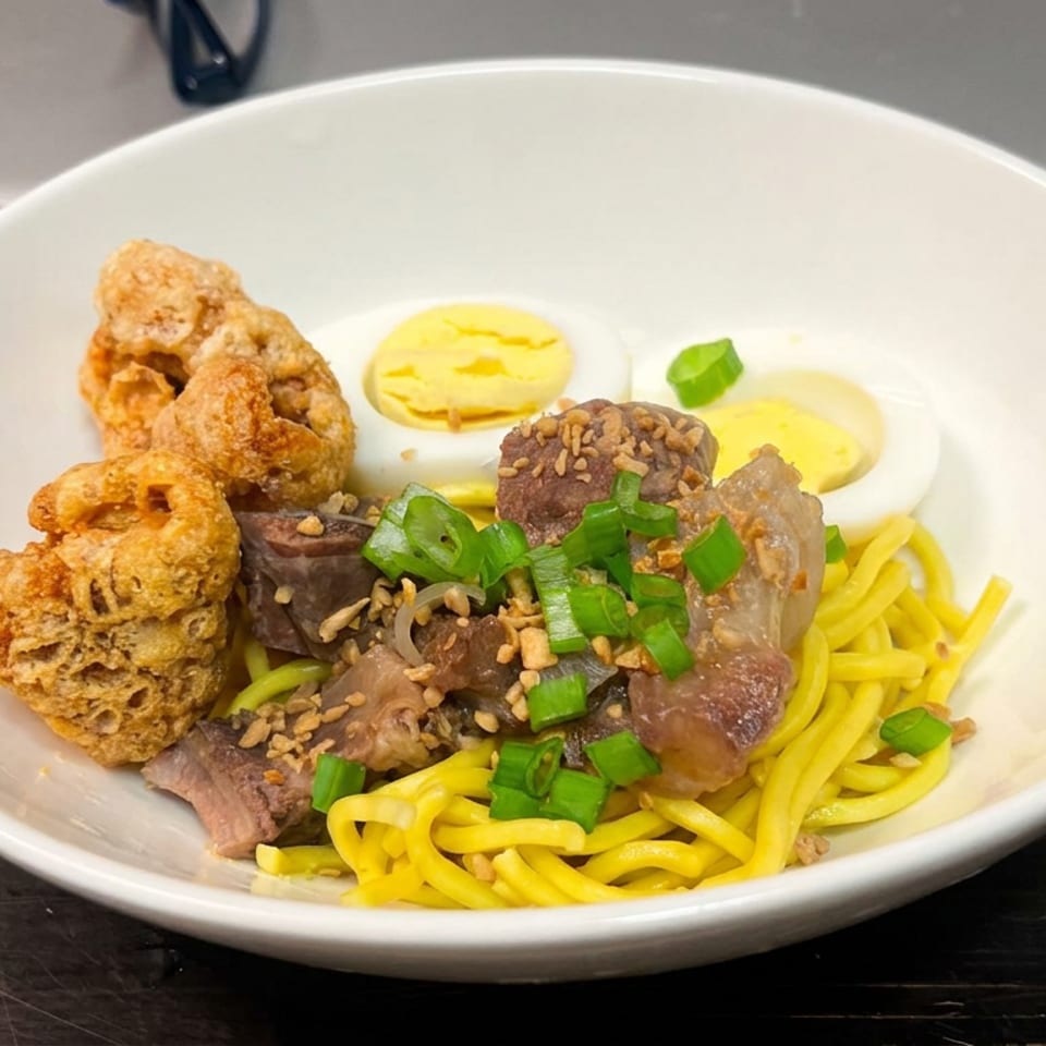 Bowl of yellow noodles topped with sliced meat, fried mushrooms, boiled egg, sesame seeds, and green onions.